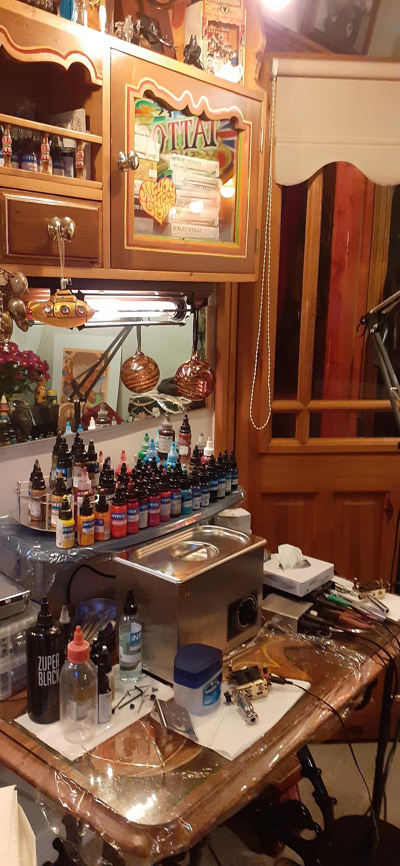 Cozy DC Tattoo Saloon studio with wooden cabinetry and wall decor. Ink bottles are organised on a counter beside various tattoo equipment and a magnifying lamp. A small window with blinds is visible in the background.