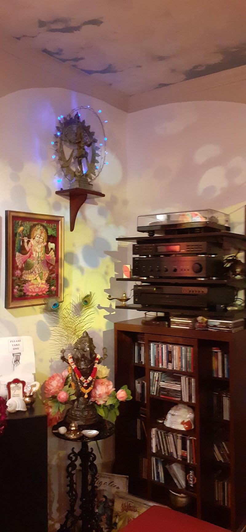 A cozy room corner featuring a music system on a wooden shelf with a stack of CDs and a stereo. Decor includes a statue adorned with lights, an artful painting reminiscent of Tattoo Nottingham, and colourful floral arrangements casting soft, patterned shadows on the walls.